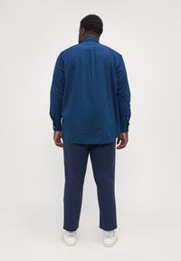 Blå skjorta med knappar och små vita prickar, långa ärmar och rundad nederkant, i kombination med mörkblå byxor och vita sneakers.