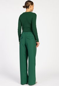 Vrouw met een bun in haar haar, gekleed in een donkergroene ribgebreide lange mouwtop en bijpassende wijde broek, staand met de rug naar de camera op een witte achtergrond.