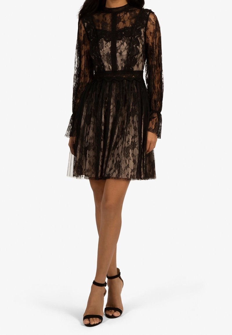 Femme portant une robe en dentelle noire longueur genoux avec manches longues et col montant, associée à des escarpins noirs à bride cheville.