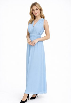 Femme debout portant une robe longue sans manches bleu clair et des talons hauts noirs, mains jointes devant, fond blanc neutre.