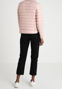 Kvinde iført en lys pink quilted jakke, sorte cropped bukser og hvide sneakers, stående med ryggen mod en ensfarvet baggrund.