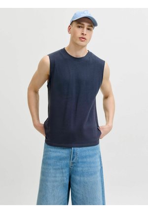 Noor mees kannab helerohelist nokamütsi, varrukateta tumesinist särki ja lahtisi helerohelisi teksapükse, seisab käed taskutes.