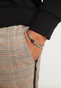 Hopeaketju, jossa on pyöreä musta aksentti, on kädessä, yhdistettynä mustaan pitkähihaiseen paitaan ja beigeihin ruudullisiin housuihin, joissa on oransseja yksityiskohtia.