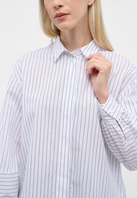 Hvid skjorte med knapper og lodrette marineblå striber, lange ærmer med knappede manchetter og en almindelig krave. Letvægtsstof med en let glans.