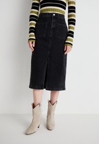 Jupe midi en denim noir avec fente avant et deux poches arrière. Portée avec des bottes de cowboy en suède beige. Un haut à rayures à manches longues est visible au-dessus.