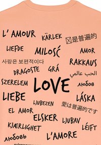 T-shirt couleur pêche avec un texte noir comportant le mot "AMOUR" et des traductions dans diverses langues, disposées de manière ludique sur le tissu.