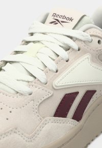 Tuvumā redzama bēša Reebok športa apava ar baltiem diegiem, bordo logotipa svītrām un teksturētām zamšādas un ādas detaļām.