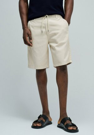 Homme portant un short beige à cordon, des sandales noires avec boucles, et une chemise bleu marine foncé, debout avec une main dans la poche.