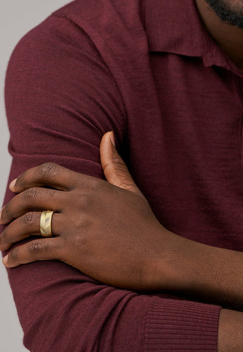 Manica di un maglione a maglia bordeaux con texture a coste, braccio incrociato sopra la mano che mostra un anello color oro con un motivo diagonale.