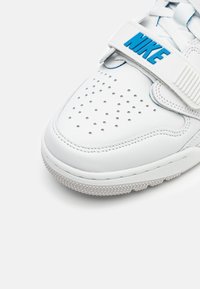 Bout de chaussure blanche Nike avec perforations, semelle texturée, et logo Nike bleu sur une sangle blanche au-dessus des lacets blancs.