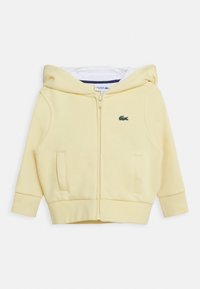 Sudadera amarilla con cremallera fabricada en material suave, con un bolsillo tipo canguro, puños de canalé y un pequeño logo verde en el pecho.