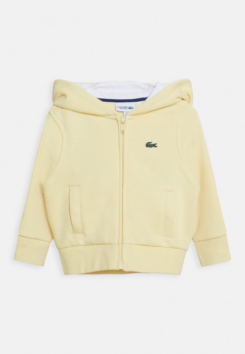 Sudadera amarilla con cremallera fabricada en material suave, con un bolsillo tipo canguro, puños de canalé y un pequeño logo verde en el pecho.
