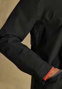 Jachetă impermeabilă neagră, cu o textură netedă, manșete elastice cu accente portocalii și buzunare frontale sigure cu închideri cu fermoar.