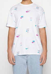 Camiseta blanca de algodón con varios logotipos de Nike en rosa y azul, y el texto "JUST DO IT" en gris, sobre un fondo liso.