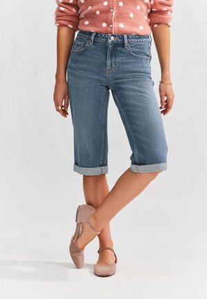 Personne portant un short en jean remonté aux genoux, un pull rose à pois et des ballerines beiges à bride à la cheville, debout jambes croisées.
