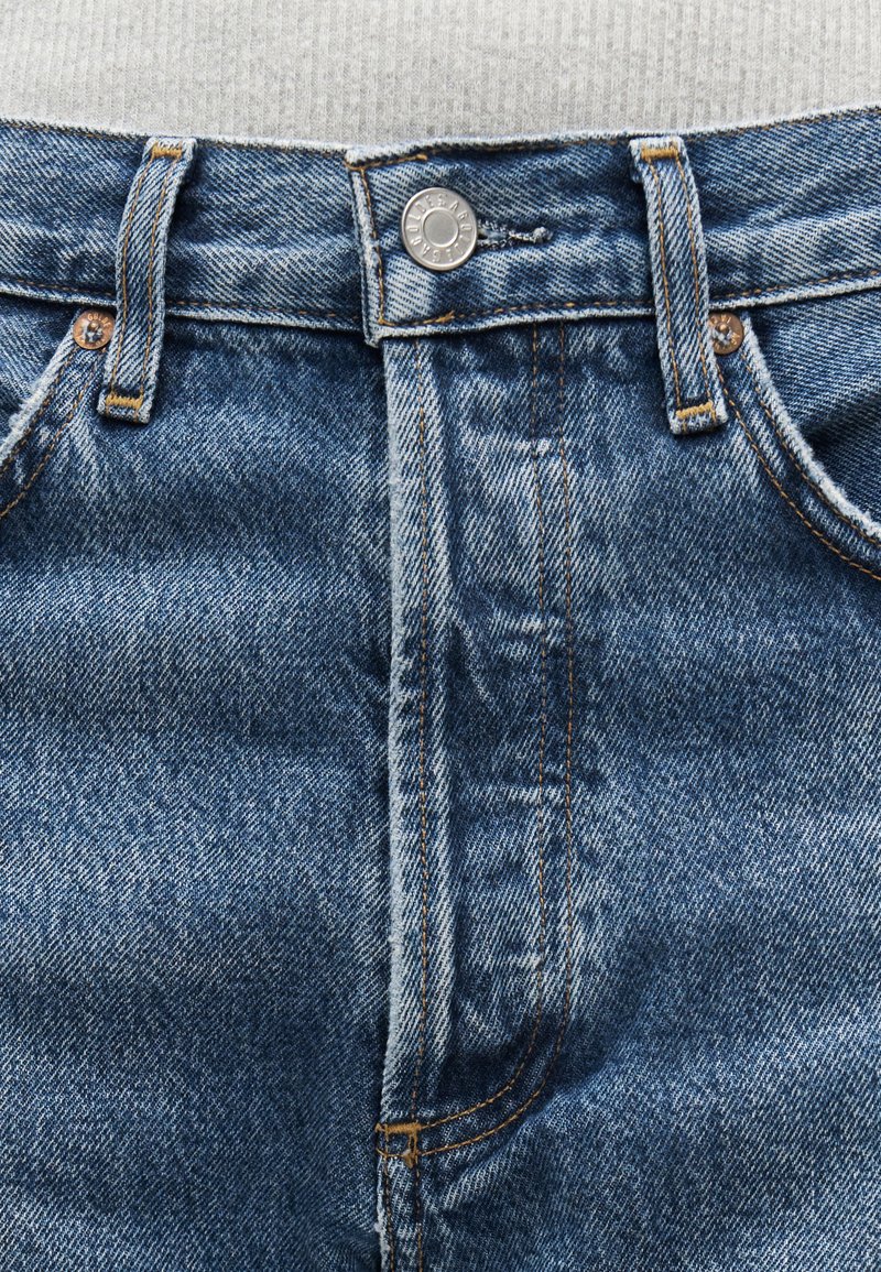 Gros plan sur la taille d'un jean en denim bleu avec un bouton en métal, des passants de ceinture, des poches avant et des détails de couture visibles.