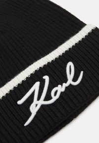 Bonnet en tricot noir à cote avec une rayure horizontale blanche et un logo "Karl" brodé. Texture douce et ajustement serré pour la chaleur.