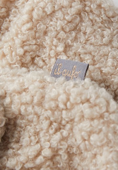 Μπεζ, αφρώδες ύφασμα με κυματιστή υφή. Περιλαμβάνει μία μικρή γκρίζα ετικέτα με τη λέξη "ilaufo" ραμμένη σε κρέμα. Μαλακή εμφάνιση.