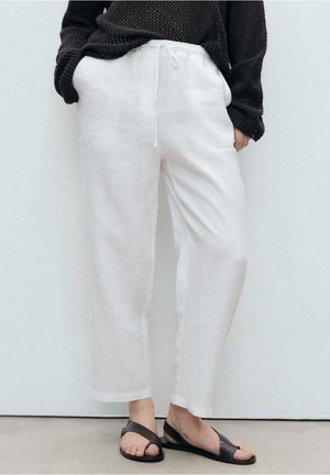 Persoon draagt loszittende witte linnen broek met trekkoord in de taille, zwarte gestructureerde trui en zwarte sandalen met open tenen, staand tegen een witte muur.