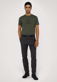 Olivově zelené tričko s černým logem, kombinované s tmavě šedými slim fit kalhotami. Černé kožené boty dokončují outfit a podtrhují jeho design.