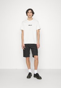 Weißes Baumwoll-T-Shirt mit schwarzem "ellesse"-Logo, schwarze Shorts und schwarze Schuhe. Das Modell trägt weiße Socken; einfaches Design, keine Muster oder Akzente.