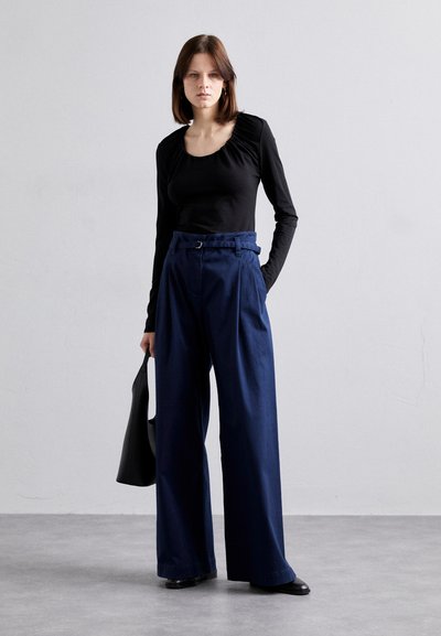 Schwarzes langärmliges Oberteil mit gerafftem Ausschnitt, kombiniert mit hoch geschnittenen, weit geschnittenen navyblauen Hosen. Das Model hält eine schwarze Tasche.