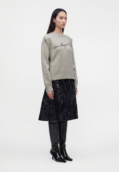 Femme portant un pull gris avec une inscription noire, une jupe noire texturée et brillante, et des bottes noires à talons montant jusqu’aux genoux, debout sur fond blanc.