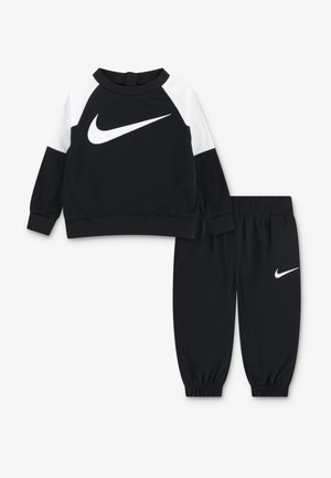 Črni komplet sestavljen iz sweatshirt-a in hlač, ki ima bel Nike logotip. Sweatshirt ima izrazite bele ramena; hlače imajo elastične manšete. Mehak material.