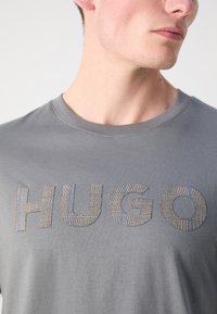 Pelēks kokvilnas T-krekls ar vārdu "HUGO" strukturētā auduma rakstā ar smalkām līnijām un zilām akcentiem. Ar apaļu kakla izgriezumu.