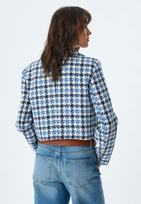 Kort jacka med ett vävt mönster i blått, svart och vitt rutmönster. Har långa ärmar och en mjuk textur, tillsammans med högmidjade jeans.