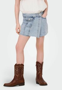 Gonna di jeans a vita alta, lavaggio azzurro chiaro, orlo sfrangiato e due tasche frontali. Abbinata a stivali da cowboy marroni con cuciture decorative.