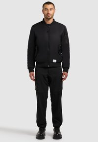 Giacca bomber nera con chiusura a zip, tasche laterali e polsini a coste. Abbinata a pantaloni cargo neri e stivali neri con lacci.