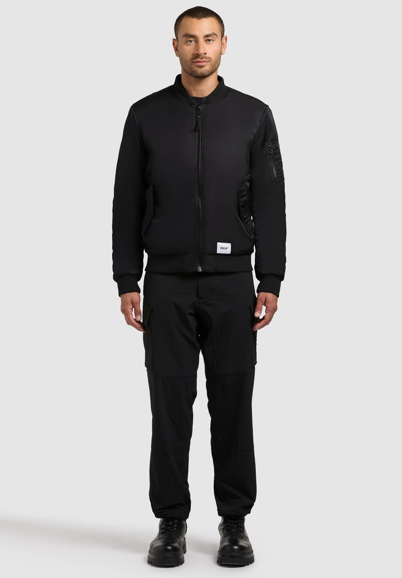 Giacca bomber nera con chiusura a zip, tasche laterali e polsini a coste. Abbinata a pantaloni cargo neri e stivali neri con lacci.