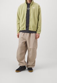 Ljust grön fleecejacka med hög krage och Nike-logotyp, kombinerat med beige baggy cargobyxor och svarta sneakers med gummisulor.