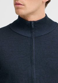 Sötét navy kék félzipzáras pulóver, magas bordázott gallérral és sima, texturált anyaggal. A cipzár mentén látható a felső varrás.