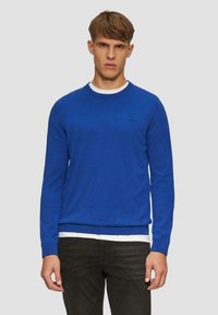 Blauer Strickpullover mit rundem Halsausschnitt und gerippten Bündchen. Verfügt über einen weißen Innenkragen und ein kleines Logo auf der Brust. Getragen mit dunkelblauen Jeans.
