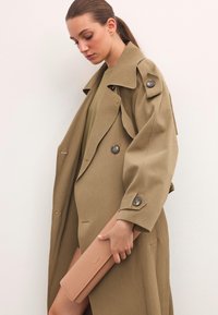 Khaki płaszcz trench z dużymi guzikami i teksturowanym wykończeniem, trzymany ręką chwytającą jasnoróżowy portfel. Prosty i elegancki design.