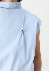 Top senza maniche di un leggero blu, con colletto alto, caratterizzato da un dettaglio di tessuto annodato e un accento decorativo in argento sul retro.