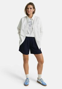 Chemise blanche à boutons sur un t-shirt graphique, associée à un short bleu marine. Sneakers bleues avec des accents marron et chaussettes blanches complètent le look.