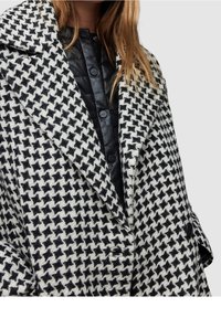 Schwarz-weißer Hahnentritt-Mantel mit Reverskragen, doppelt geknöpftem Vorderteil und strukturiertem Stoff; darüber ein schwarzes Hemd mit Knöpfen getragen.