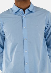 Camisa azul clara con un patrón texturizado, que presenta un cuello clásico, cierre de botones y un logo blanco en el pecho. Tela arrugada.