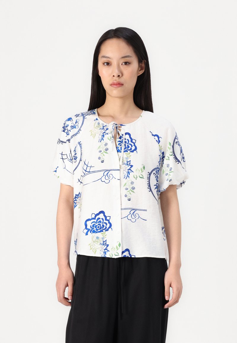Blusa bianca a maniche corte con motivi floreali blu e accenti verdi, dotata di scollatura rotonda e chiusura frontale a bottoni.