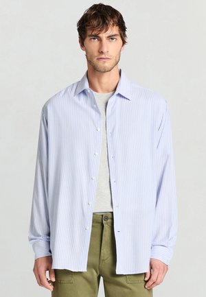 Hellblau-weiß gestreiftes Hemd aus Baumwolle mit Knöpfen, ausgestattet mit spitzem Kragen und langen Ärmeln, getragen über einem grauen T-Shirt.