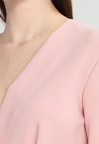 Blouse rose avec un col en V, faite d'un tissu lisse. Présente une texture subtile et une coupe décontractée, avec un joli drapé au niveau de l'épaule.