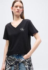 Νεαρή γυναίκα με μακριά μαλλιά, που φοράει μαύρο T-shirt Calvin Klein με V λαιμόκοψη, μπλε τζιν και μια δεμένη μαύρο-λευκή καρό πουκάμισο γύρω από τη μέση της.