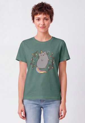 Femme aux cheveux courts et bruns porte un t-shirt vert avec une licorne chat gris à la queue arc-en-ciel entourée de délicates branches florales.