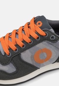 Harmaat lenkkarit, joissa on oranssit nauhat ja logo, valmistettu yhdistelmästä teksturoitua mokkaa ja sileää kangasta. Matala malli, jossa on pyöreä kärki.