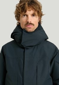 Tumma, eristetty takki, jossa on korkea kaulus ja joka näyttää sileältä ja minimalistiselta. Kangas vaikuttaa kestävältä ja tyylikkäältä.