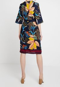 Blommönstrad klänning med trumpetärmar, en figursydd midja och kjol som når knäet. Har livfulla färger på en marinblå bakgrund.