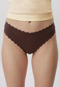 Gros plan sur une personne portant une culotte marron sans coutures à bord ondulé et un haut beige clair, visible de la taille jusqu'à mi-cuisse.